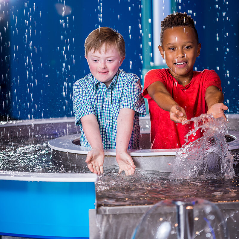 Two boys catching water as it falls from above them in the water play area.