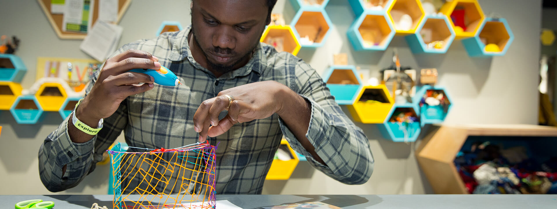 A person is crafting with a glue gun, focused on a colorful wire structure, surrounded by hexagonal shelves filled with materials.