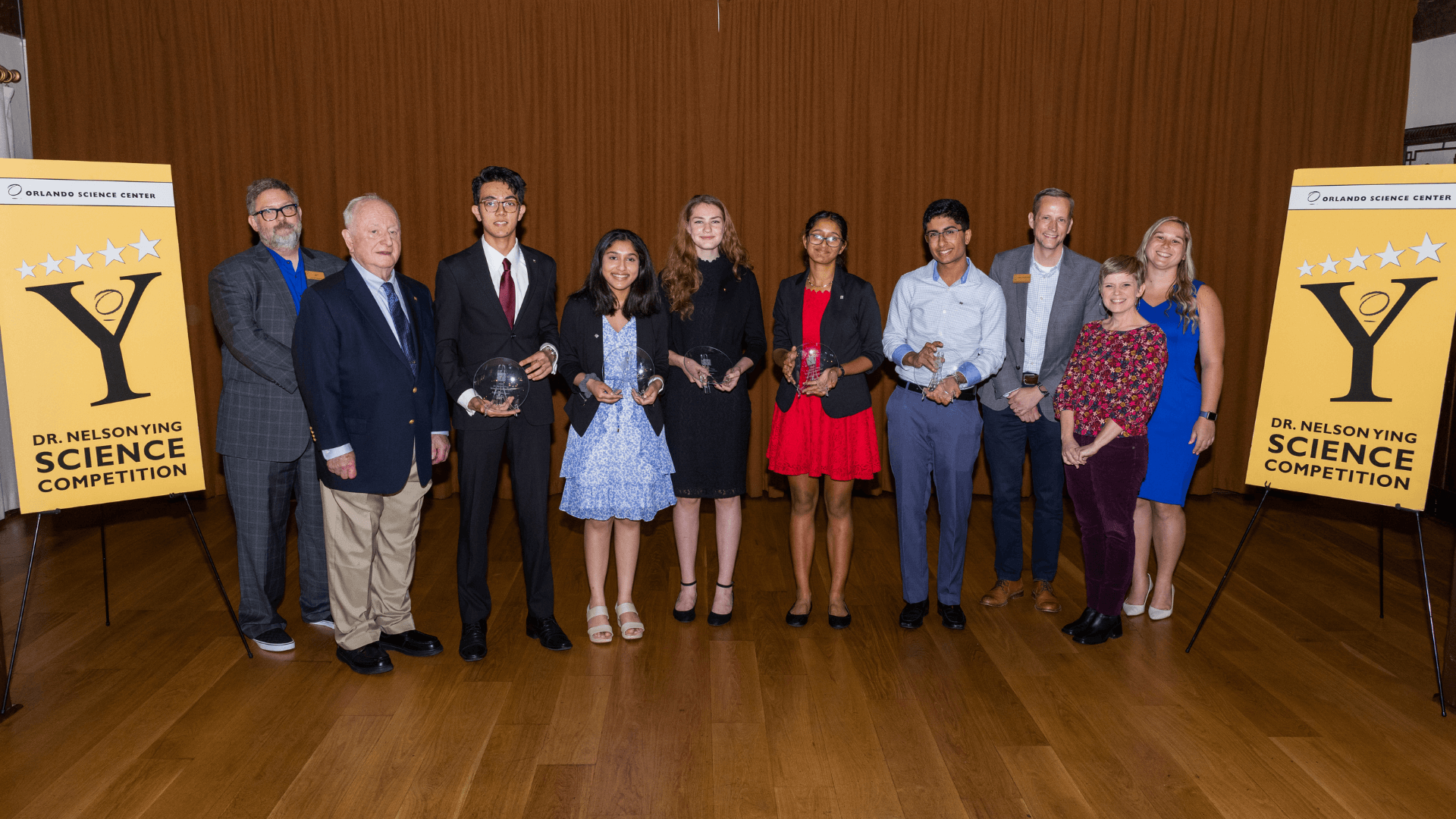 A group photo of 5 teen science competition finalists holding trophies with competition judges and OSC staff