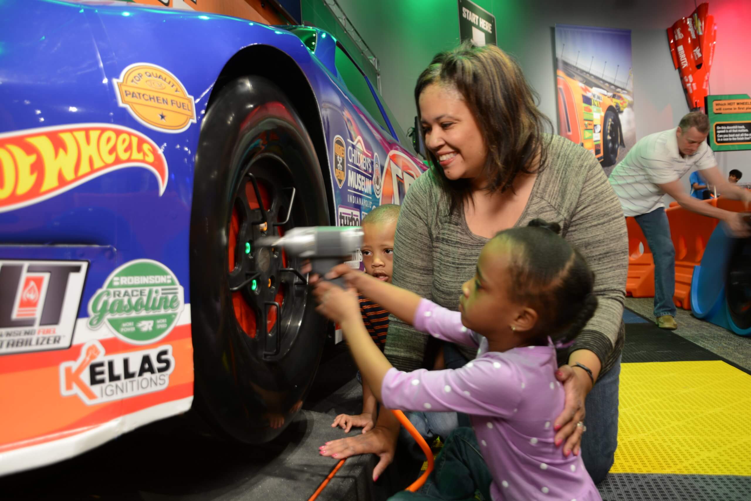A mom helps her daughter screw in a hubcap on a life-sized hot wheels race car.