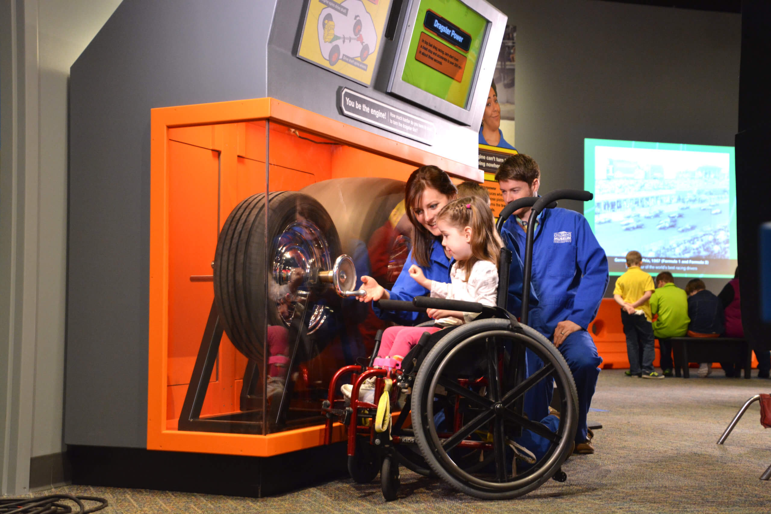 A young girl in a wheelchair turns a wheel to power a race car wheel.