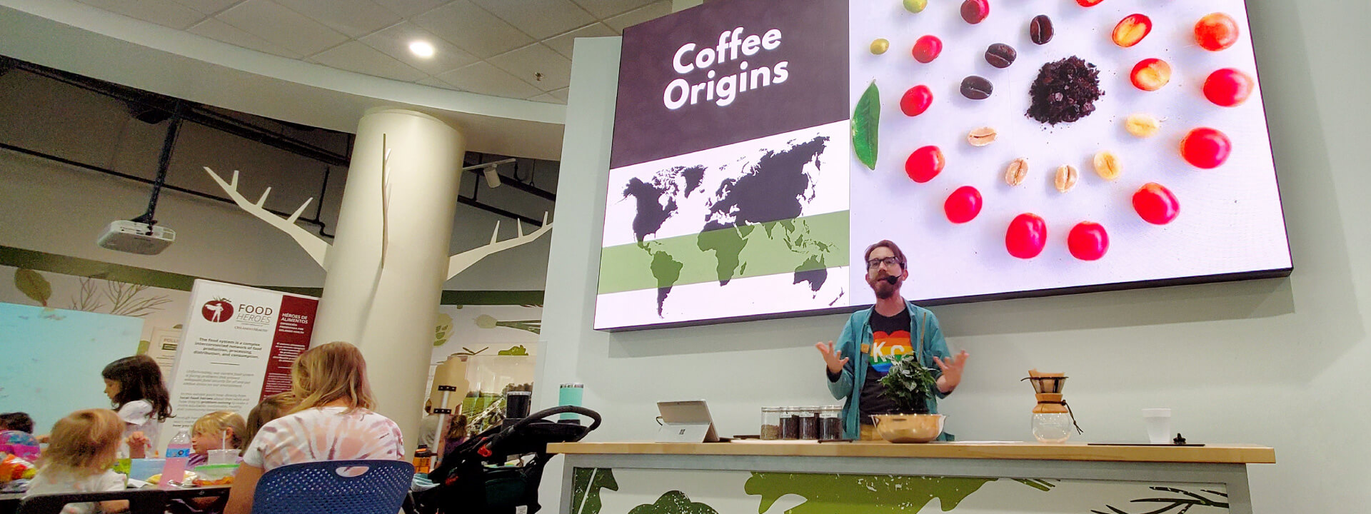 Photo of presenter giving a food science demonstration in front of large video screen in Food Heroes exhibit.