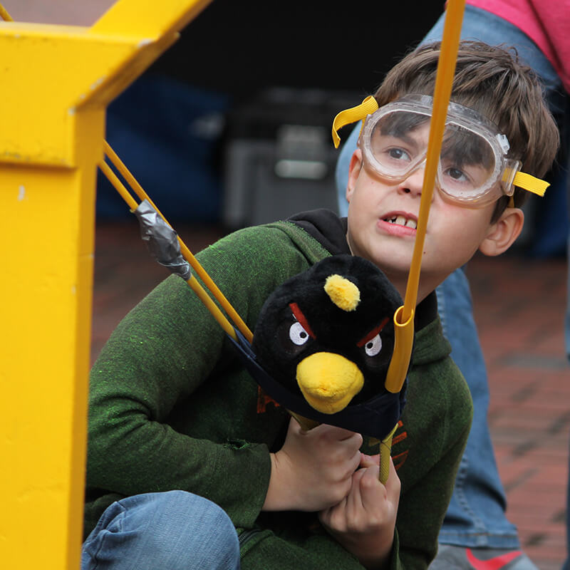 A boy wearing goggles holds a plush black bird while preparing for a launch with a slingshot setup.