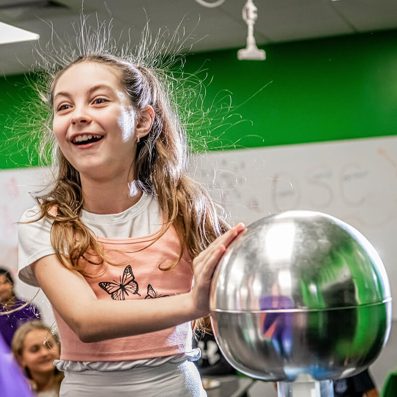 A girl with static hair smiles while touching a shiny metal sphere in a colorful classroom setting.