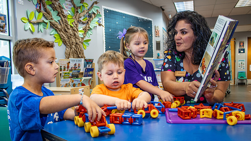 Preschool educator reading to three preschool students at a table in classroom.