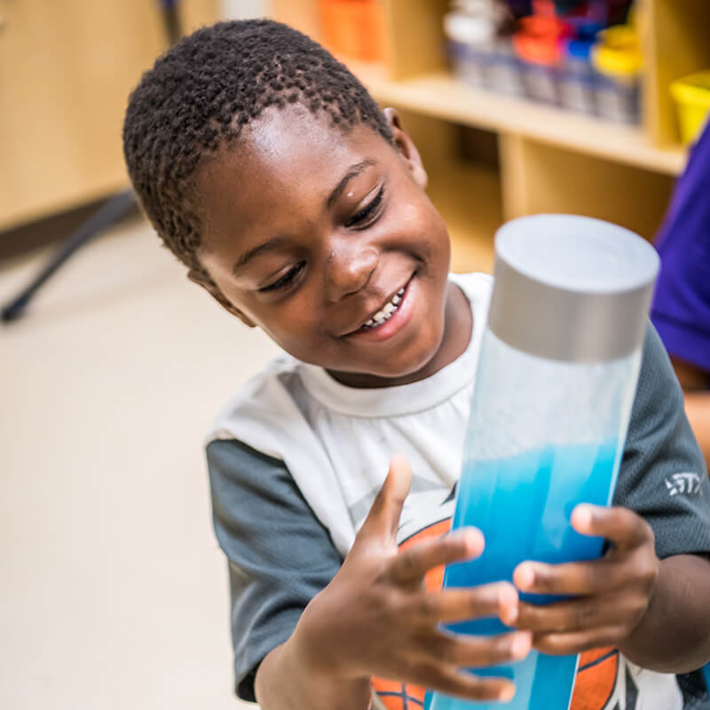 A smiling child holds a clear container filled with blue liquid, showing excitement and curiosity in a classroom setting.