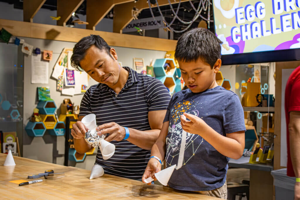 A man and a boy are engaged in a hands-on activity, constructing a project for an "Egg Drop Challenge" at a creative space.