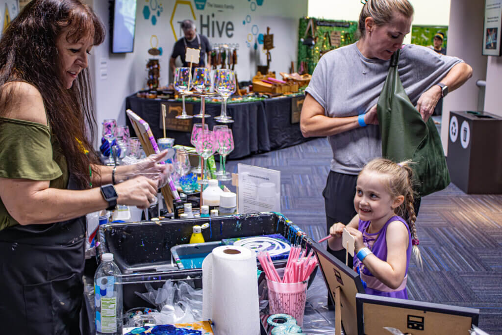 A woman assists a young girl at a craft table, surrounded by art supplies and colorful decorations. Another adult is in the background.