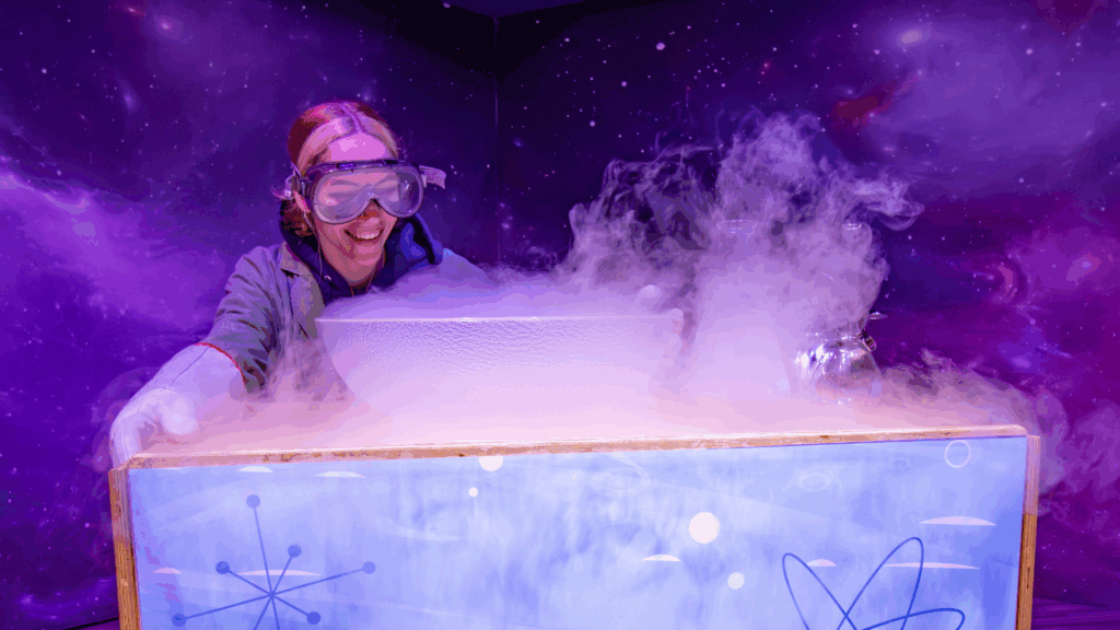 A young person in safety goggles smiles over a bowl of liquid nitrogen steam