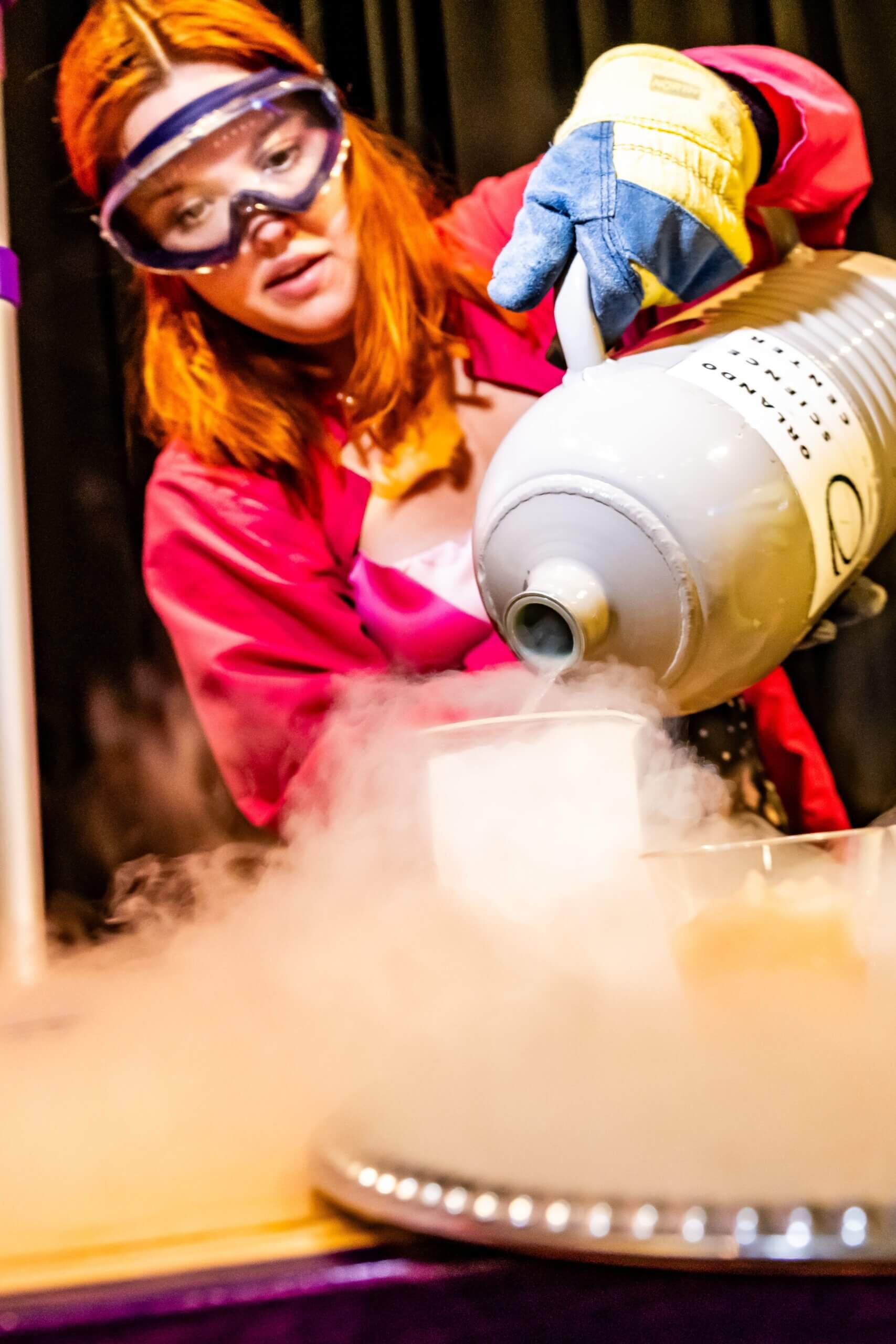 A woman in a lab coat and safety goggles pours liquid nitrogen into a bowl