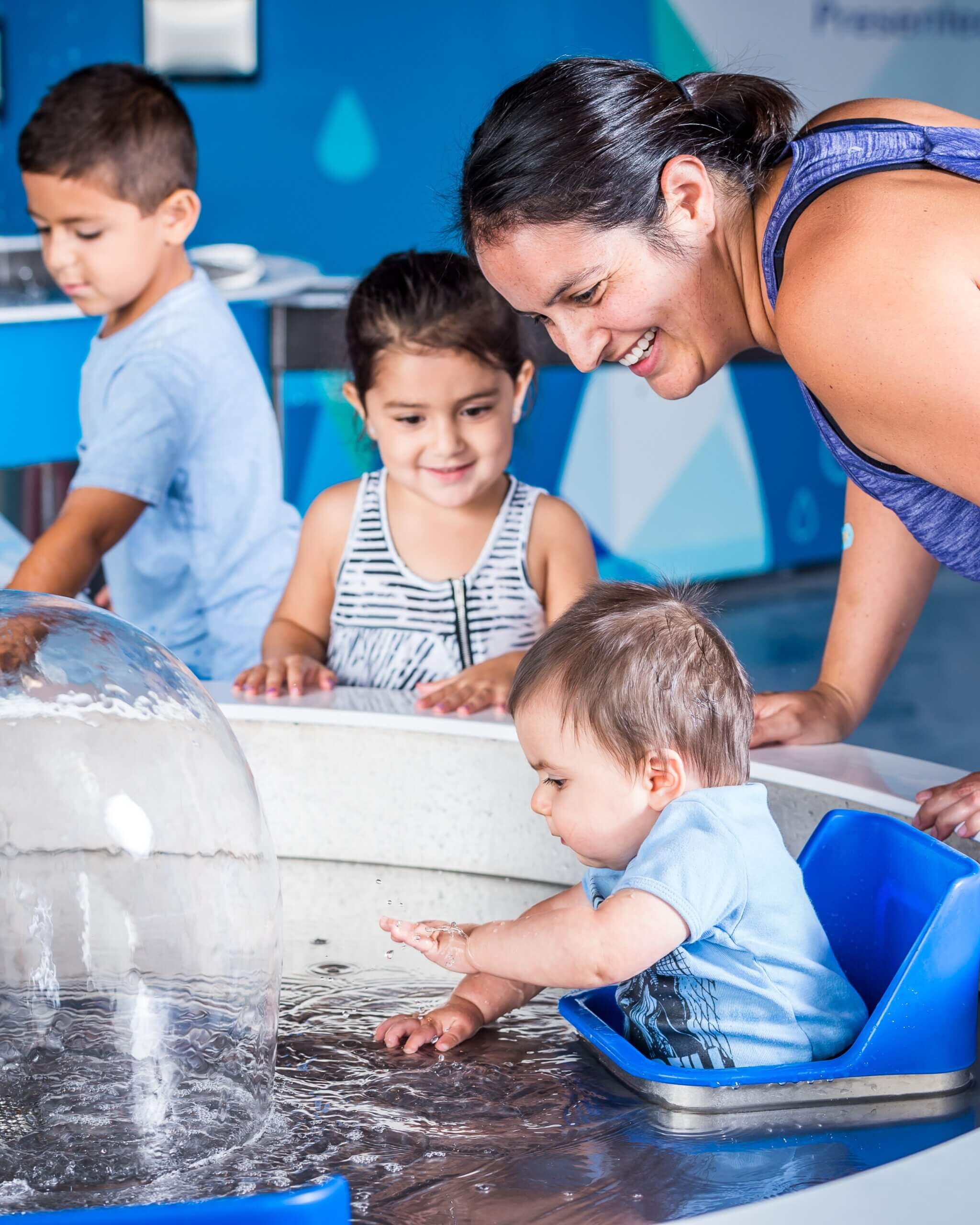 A mother and her young daughter watch a baby boy play in a water pool